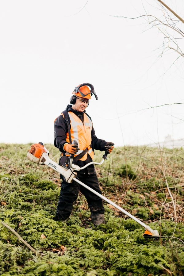 Een groenvoorziener die in de natuur staat. Hij staat in een park/bos waar geen huizen in de omgeving staan. Hij draagt werkkleding en een helm en is bezig met een machine in het gras/mos.