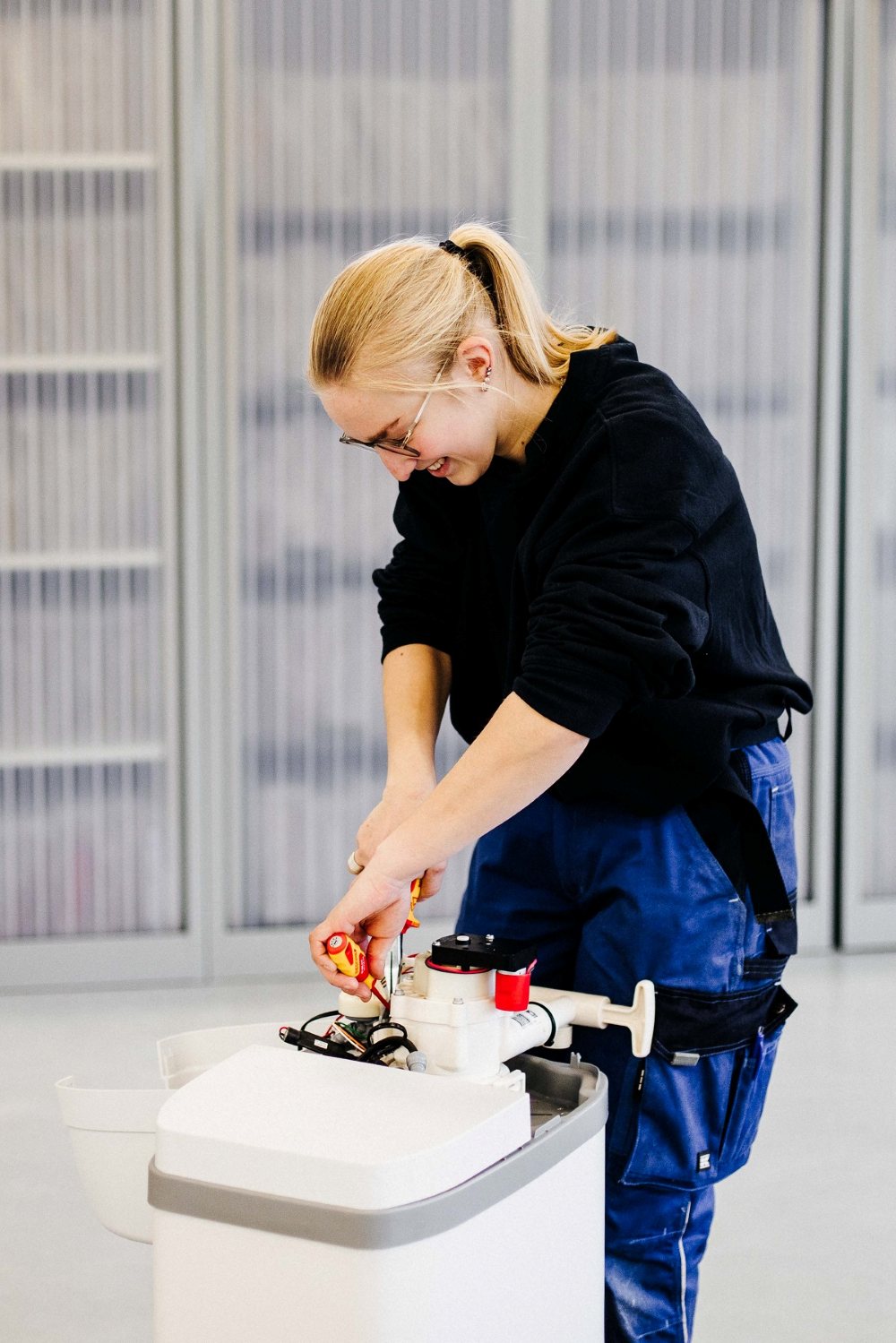 Een vrouw die bezig is met installatietechniek. Ze is iets aan het monteren en te sleutelen. Ze draagt werkkleding en lacht