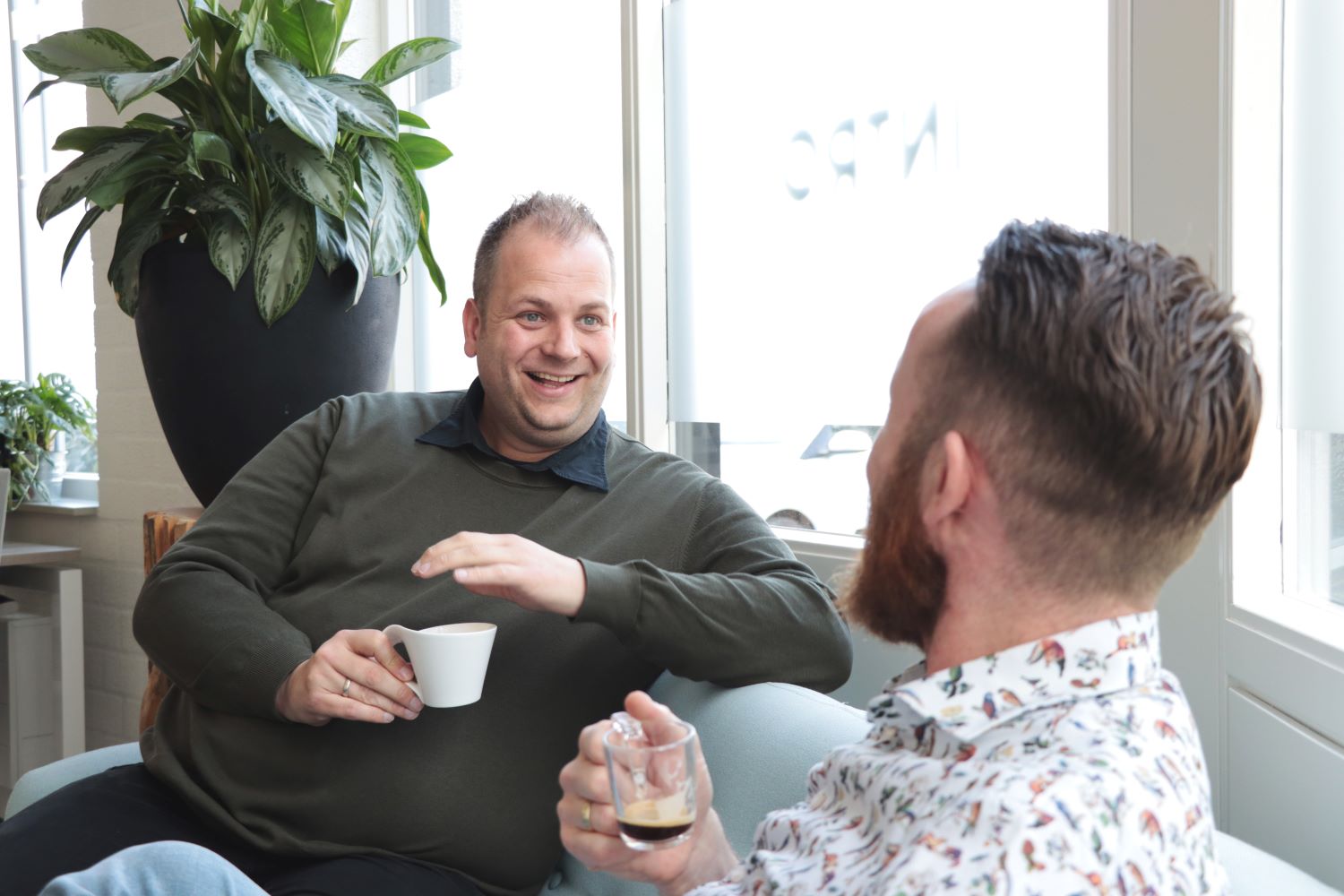 twee mannen zijn op kantoor een bakje koffie aan het drinken en ondertussen aan het overleggen.